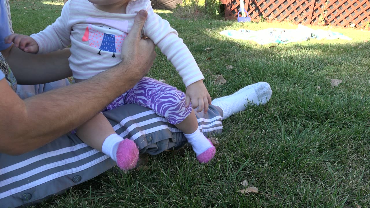 Adult steadies a toddler sitting on grass.