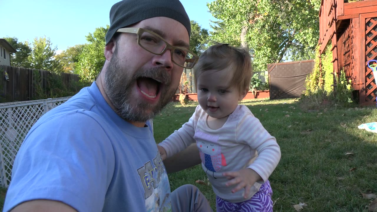 At 0:00, a bearded man in glasses and a dark beanie holds a young child close to the camera in a Westminster, Colorado backyard with grass, trees, fencing, and red wooden lattice behind them.