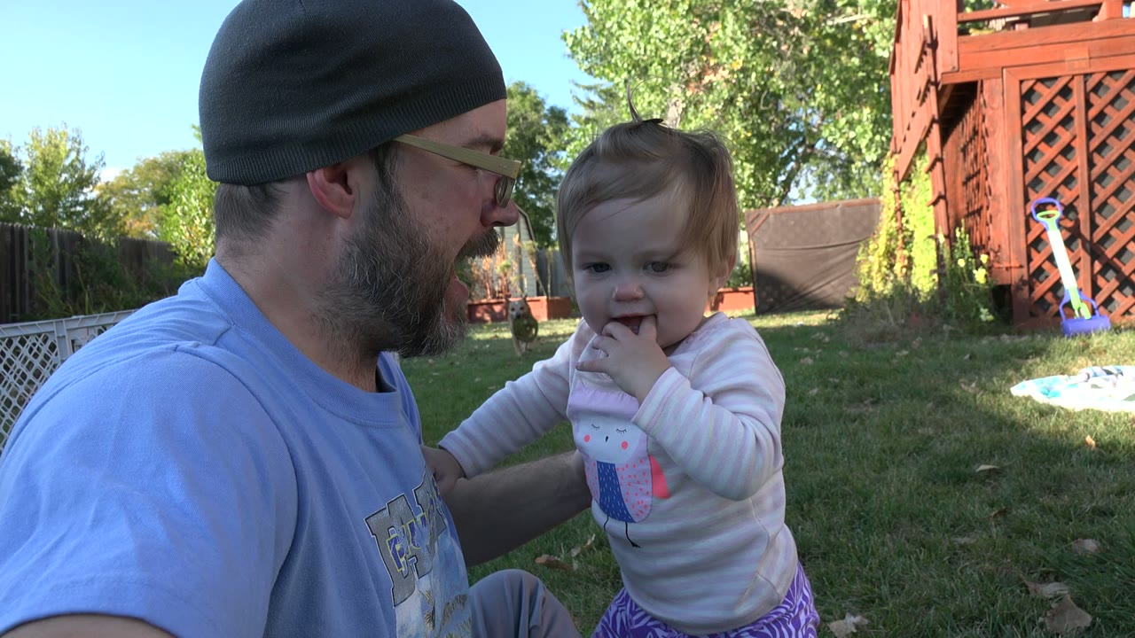 At 0:01, the man turns toward the child as she stands with a hand near her mouth in the Westminster backyard, with lawn, fence, trees, and a red wooden structure in the background.