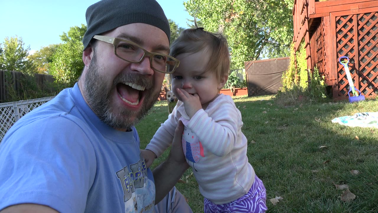 Smiling man holds a young child near his shoulder outdoors.