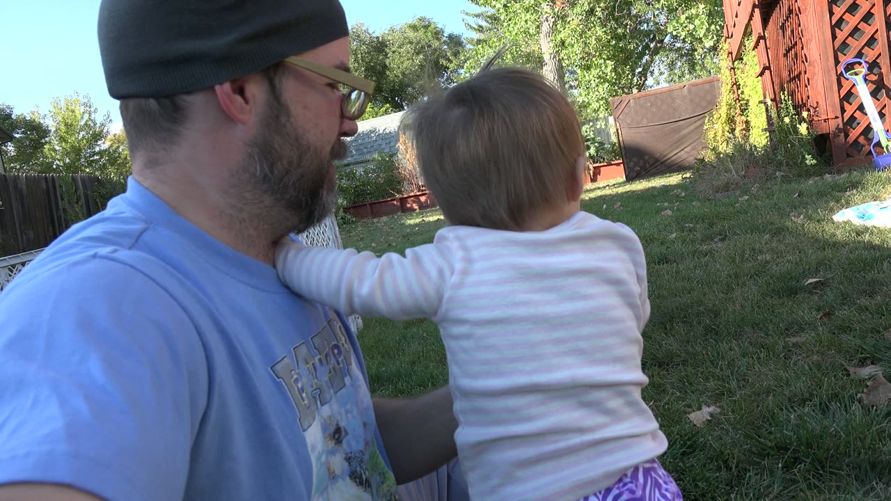Man sitting on grass with a young child facing away in a backyard.