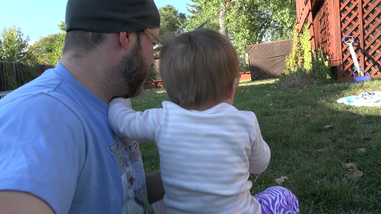In Westminster, Colorado, the man leans toward the child as they sit close together on the lawn, with a fence, trees, red lattice, and yard items behind them.