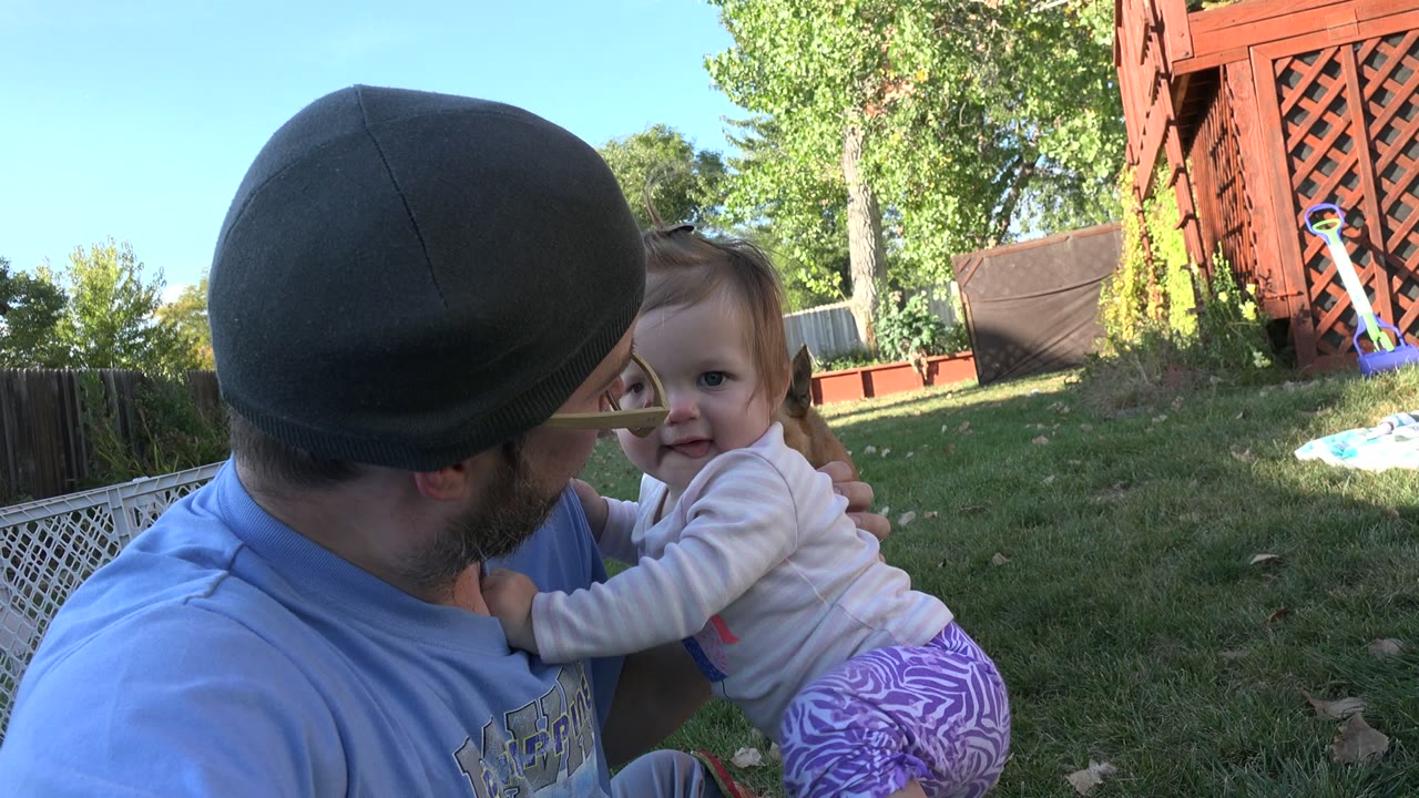 In Westminster, Colorado, a bearded man in a dark cap holds a small child close on the lawn beside a fence and red wooden yard structure.