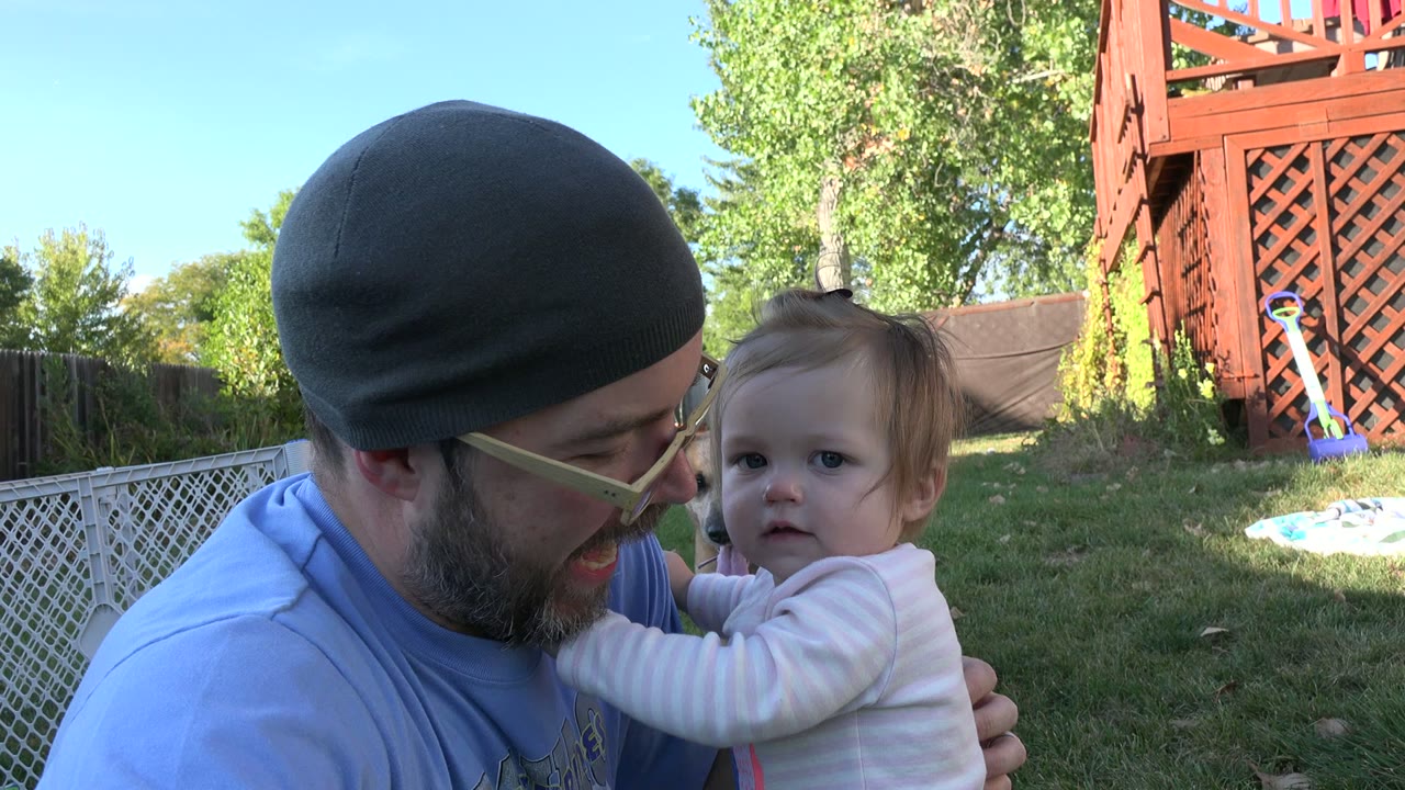 In Westminster, Colorado, the man smiles toward the child while holding them in a sunny backyard with trees, fencing, and yard items behind them.