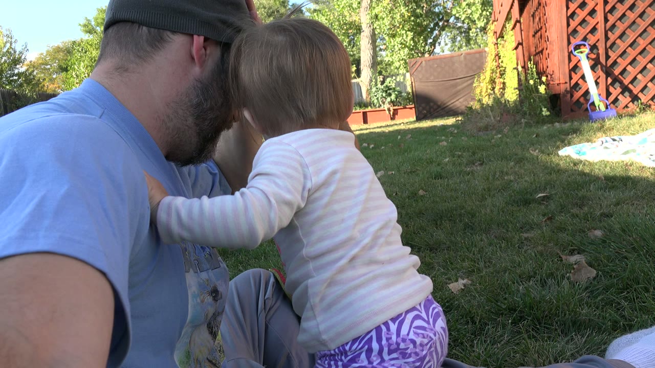 In Westminster, Colorado, the child presses close to the seated man at ground level, with sunlit grass, trees, a fence, and red lattice behind them.