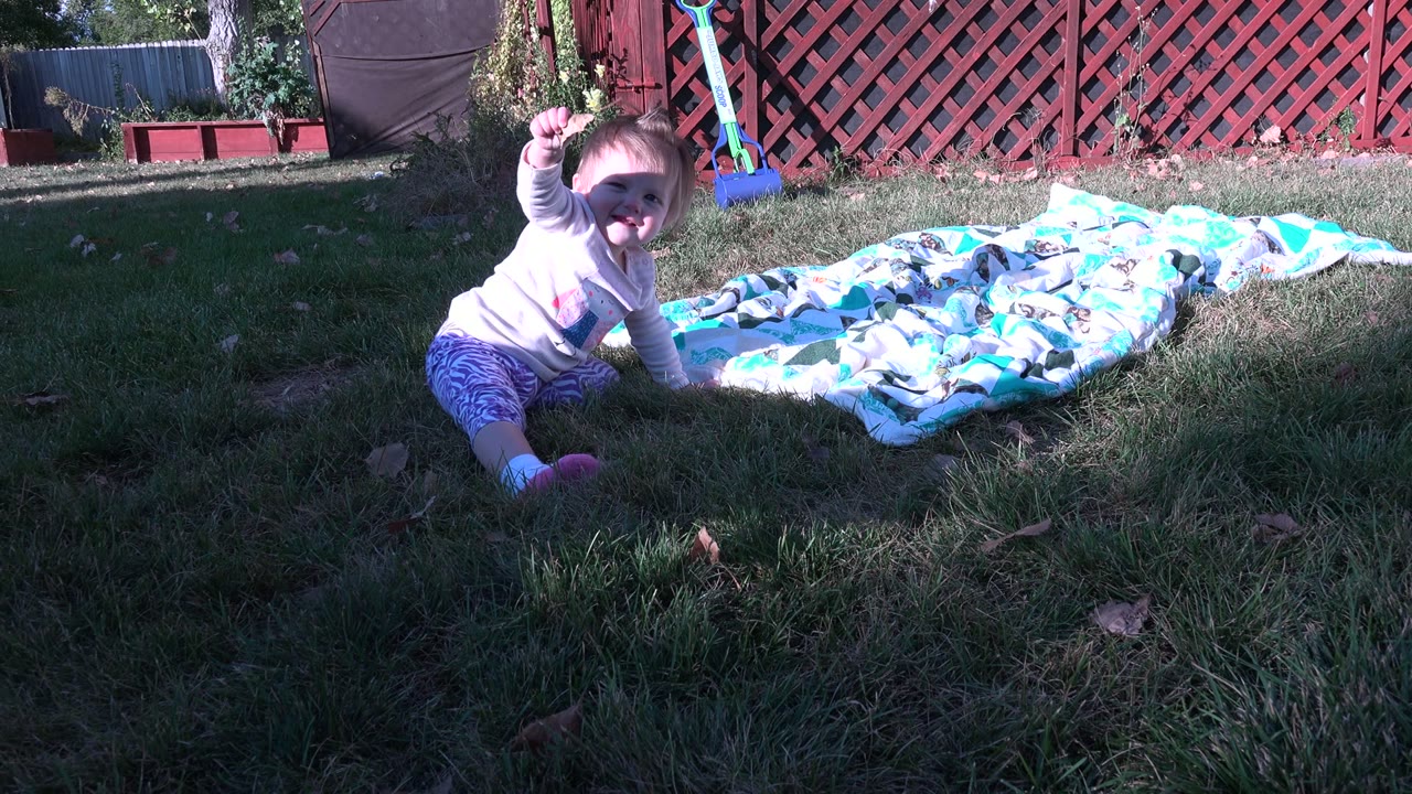 Baby sitting on grass beside a patterned blanket in a backyard.