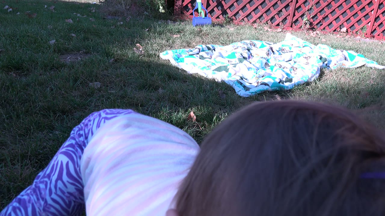 The child is bent forward near the lower center of the Westminster, Colorado backyard frame, with purple patterned pants, grass, a blanket, and red lattice fence visible.