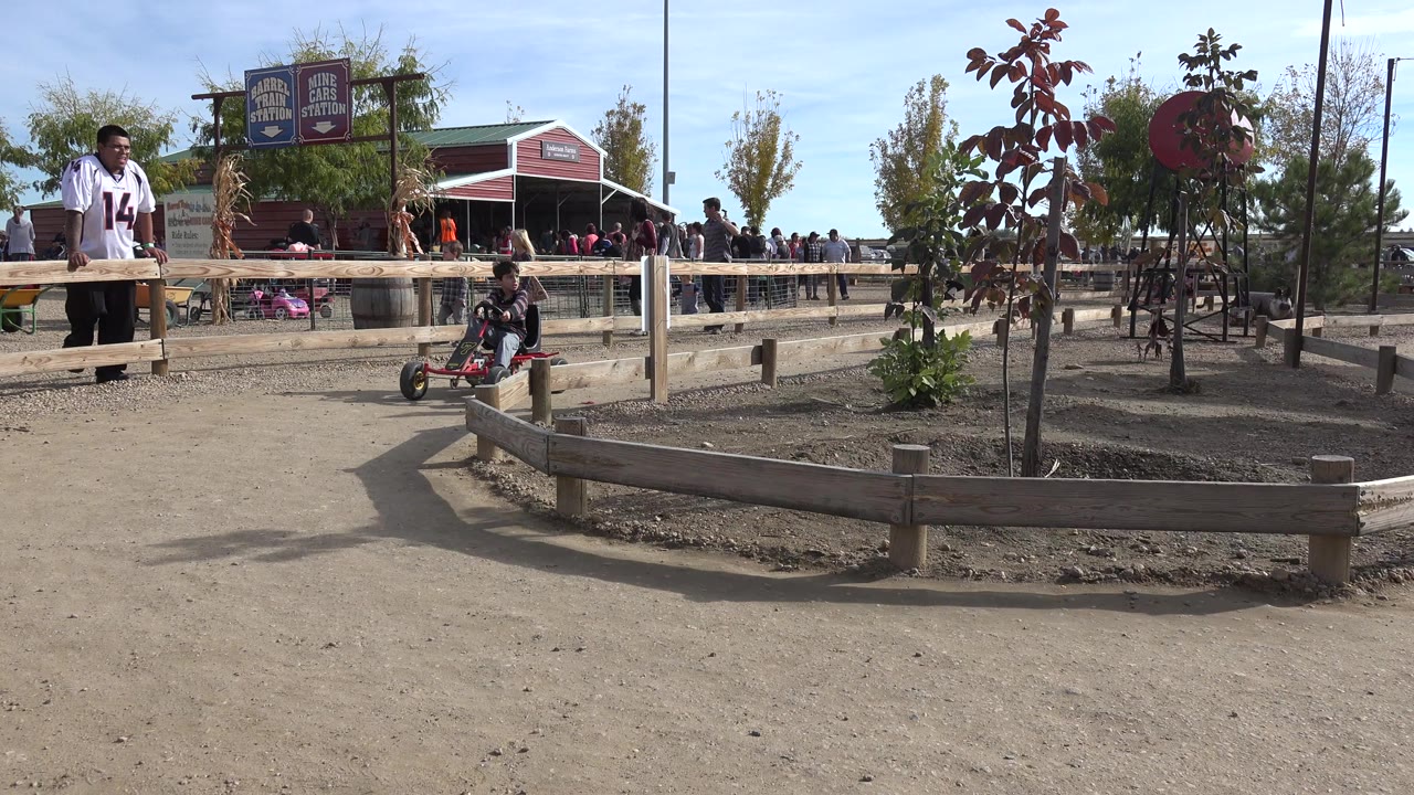 In Brighton, Colorado, a young boy on a red pedal cart rides in the midground while an adult man stands near a wooden rail and visitors gather by a red building.