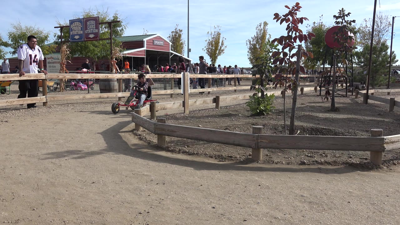 In Brighton, Colorado, the boy continues along the dirt track on a red four-wheeled pedal cart with low fencing, small trees, signs, and a red building behind him.