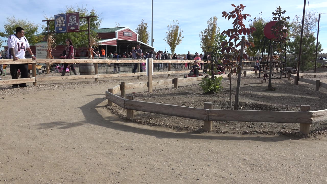 Wide park path with adults, children, wooden rail, signs, and a red-roofed building.
