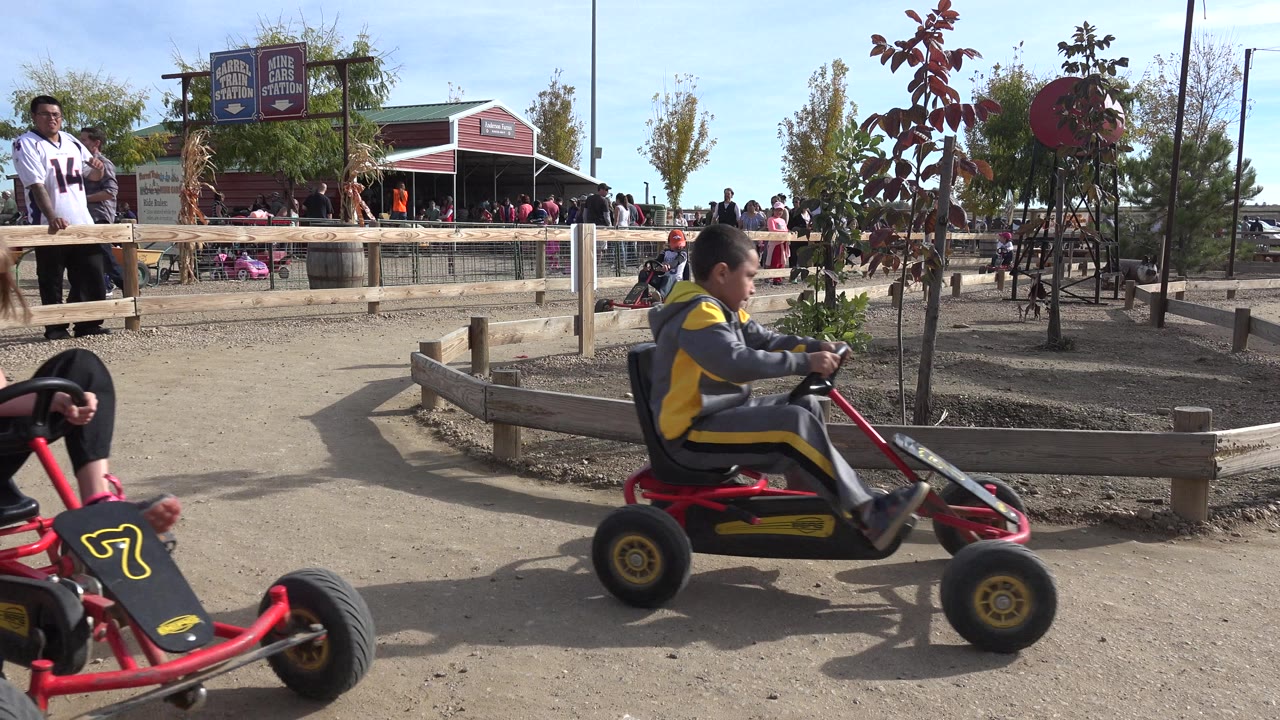 A child drives a red pedal go-kart past a low wooden fence on a dirt track in Brighton, Colorado, with adults, children, signs, and a red building in the background.