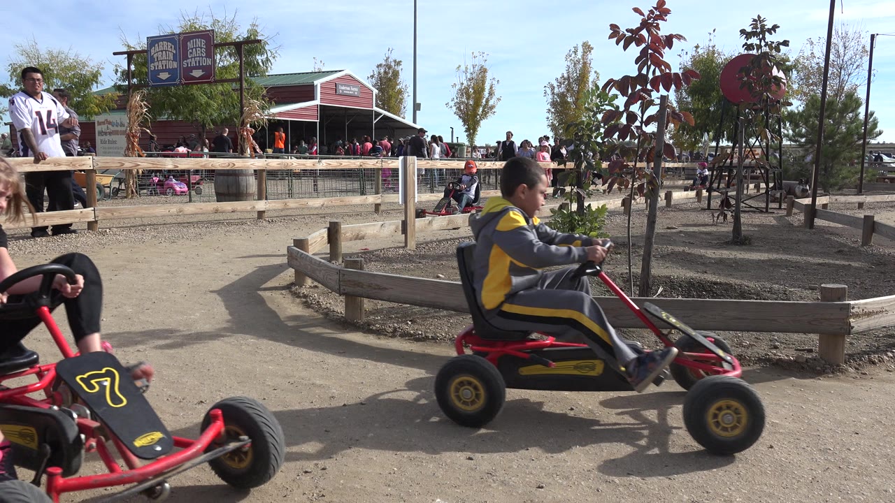 A red pedal go-kart moves close across the foreground while another child rides near the fence on the Brighton, Colorado dirt track, with spectators gathered near the red building.