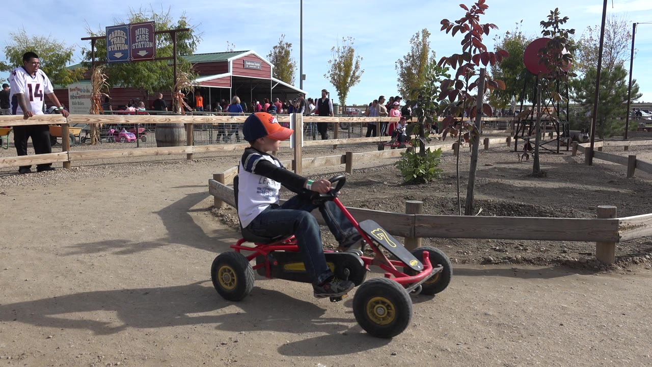 Helmeted rider on a red pedal cart near a fence and park crowd.