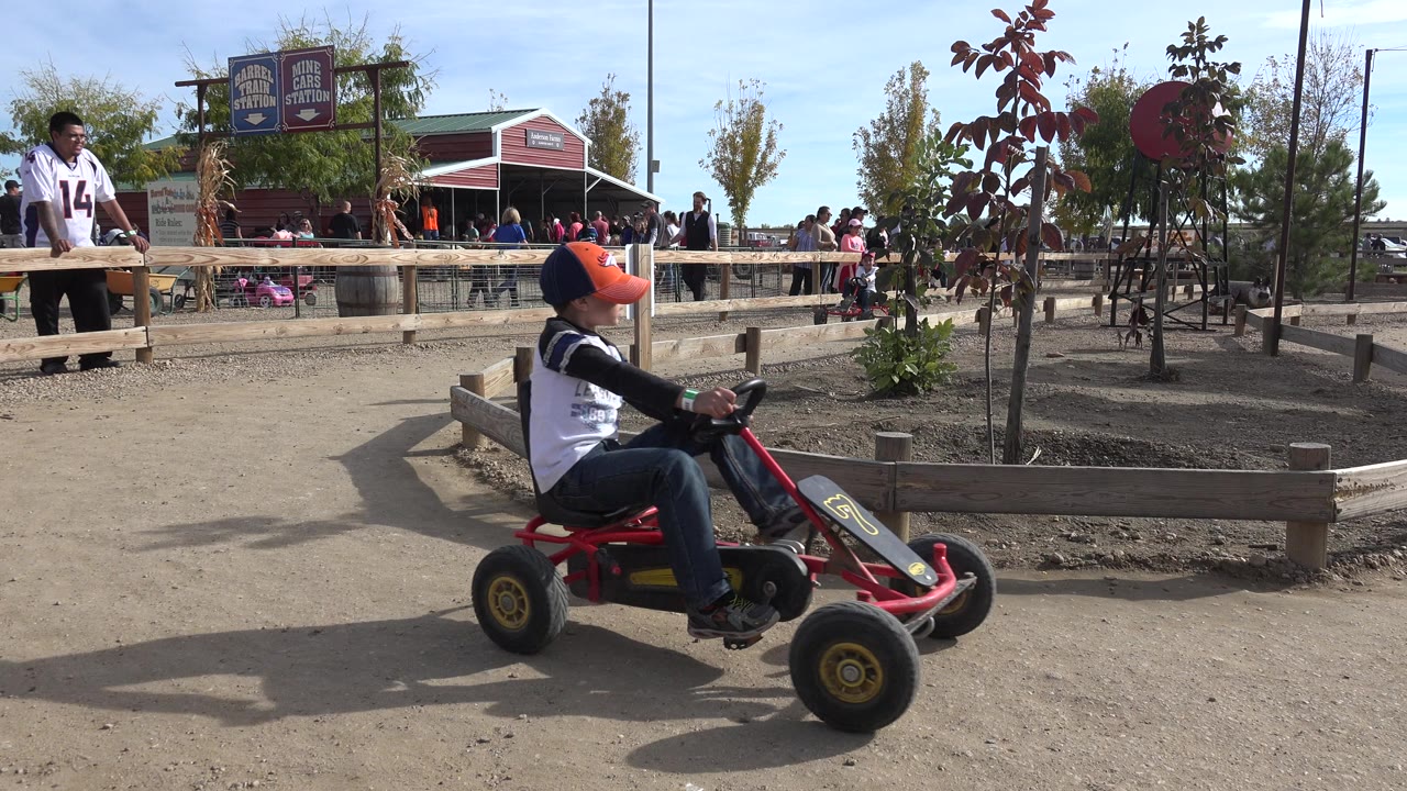 One second in, the red pedal cart is centered beside a low wooden planter fence, with Brighton park visitors and signs behind it.