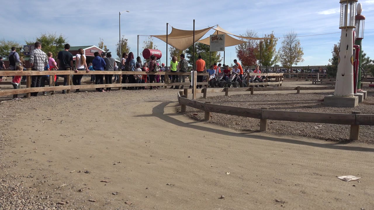 Wide low view of a Brighton, Colorado outdoor track with dusty foreground, wooden rails, shade sails, spectators, trees, and children visible near the activity area.