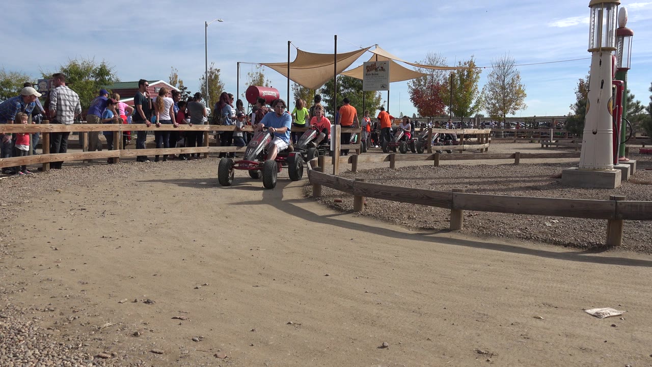 An adult rides a low four-wheeled pedal cart on a dirt path in Brighton, Colorado, with people watching behind a wooden rail and shade sails in the background.