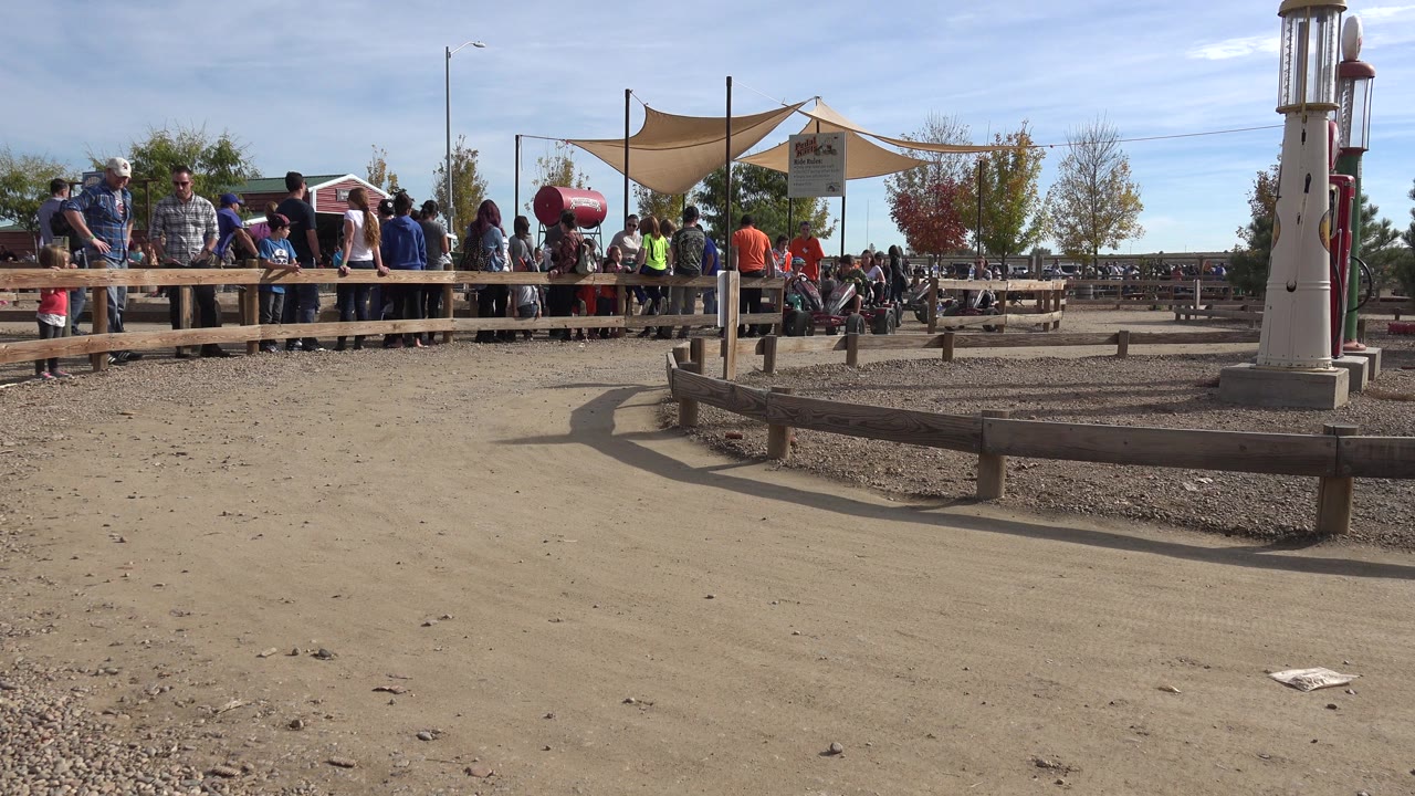 Opening frame of a Brighton, Colorado kart race start area with spectators behind a wooden rail, red karts near shade sails, and bright sun on dusty ground.