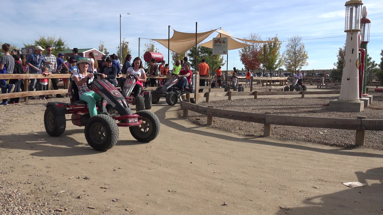 A child in a helmet rides a red pedal go-kart on a dirt track in Brighton, Colorado, with spectators behind a wooden rail and a tower structure in the background.