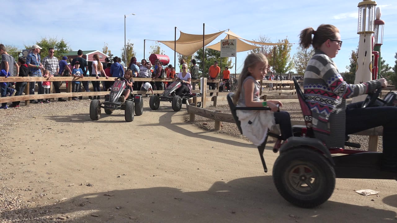 In Brighton, Colorado, the same pedal cart continues across the foreground as people watch from behind the rail fence under shade sails.
