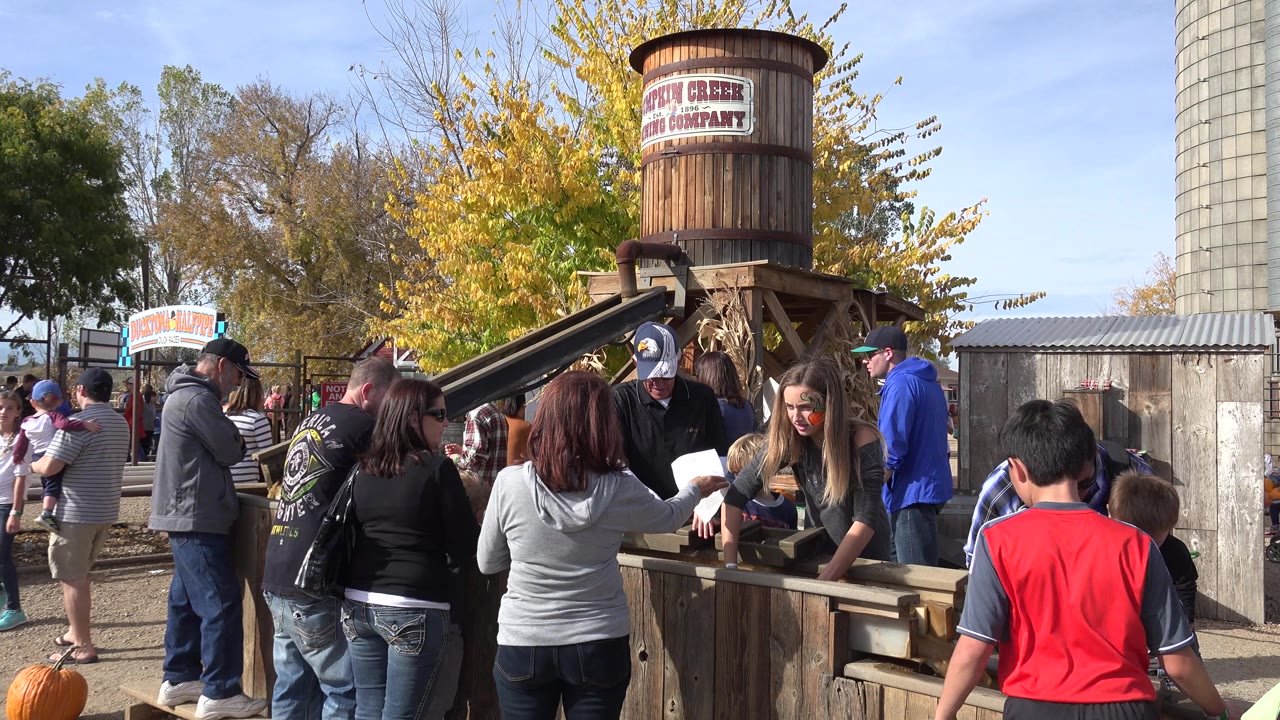 People remain packed around the wooden booth in Brighton, Colorado, while a woman reaches toward the table and children stand in the foreground under clear daylight.