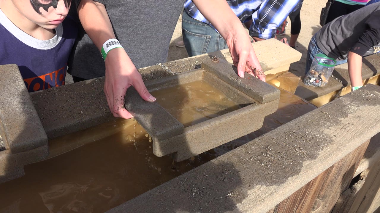 In Brighton, Colorado, a boy in sunglasses and face paint stands beside adult hands lowering a sifting tray into a muddy wooden water trough.
