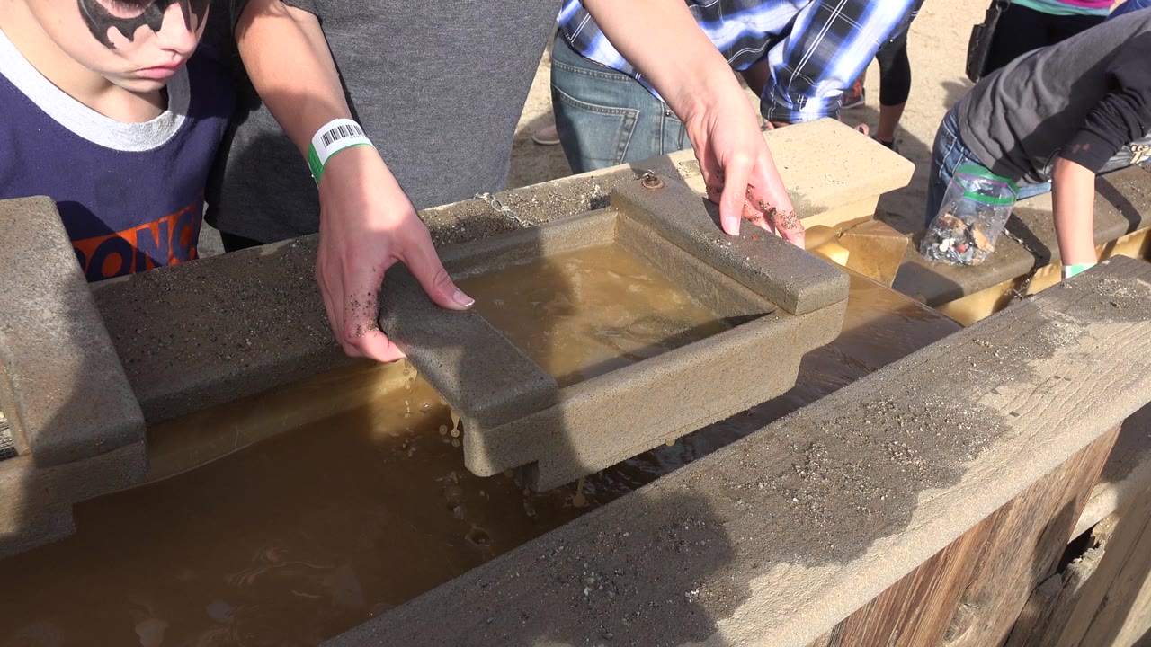 In Brighton, Colorado, hands hold a rectangular sifting tray at the edge of a muddy trough while a child in sunglasses watches from the side.