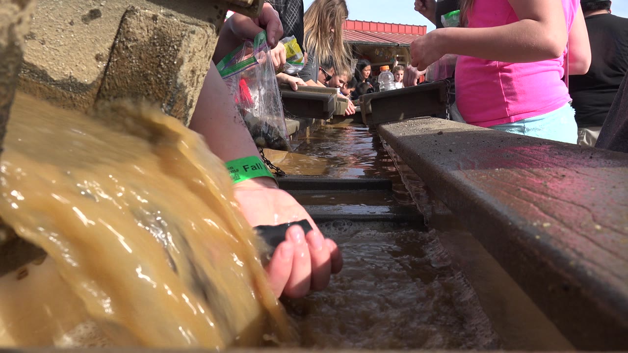 Muddy water splashes across the foreground as a hand with a green wristband rests near the flow and people stand around the Brighton trough behind it.