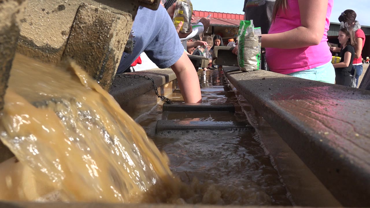At 0:00 in Brighton, Colorado, muddy water fills the foreground channel while partially visible people stand and work around trays in bright sunlight.