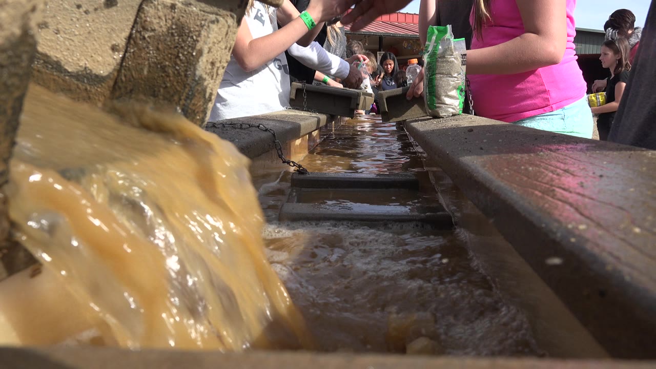 At 0:01, the muddy water stream fills the left foreground, with a wet wooden channel leading back toward sunlit people gathered around the work area in Brighton, Colorado.
