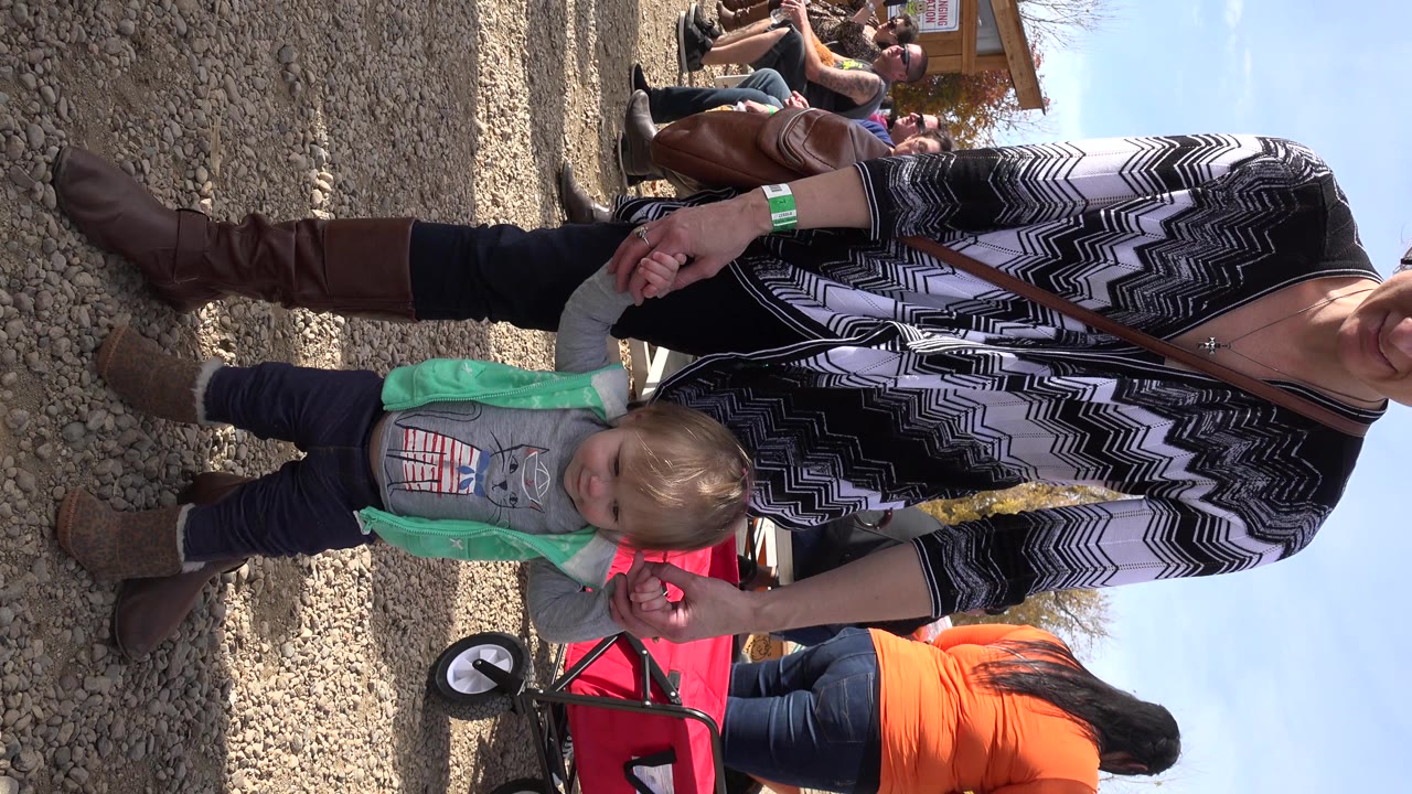 In Brighton, Colorado, the toddler remains upright on the gravel while the adult woman's hands brace him, with the red wagon wheel visible behind his shoulder.
