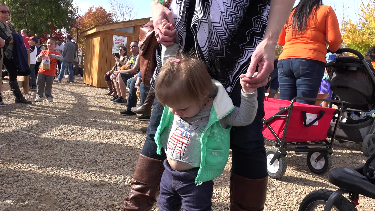 The toddler remains in the foreground in Brighton, Colorado, with an adult’s hands supporting her while a red wagon and stroller sit behind her on the gravel.