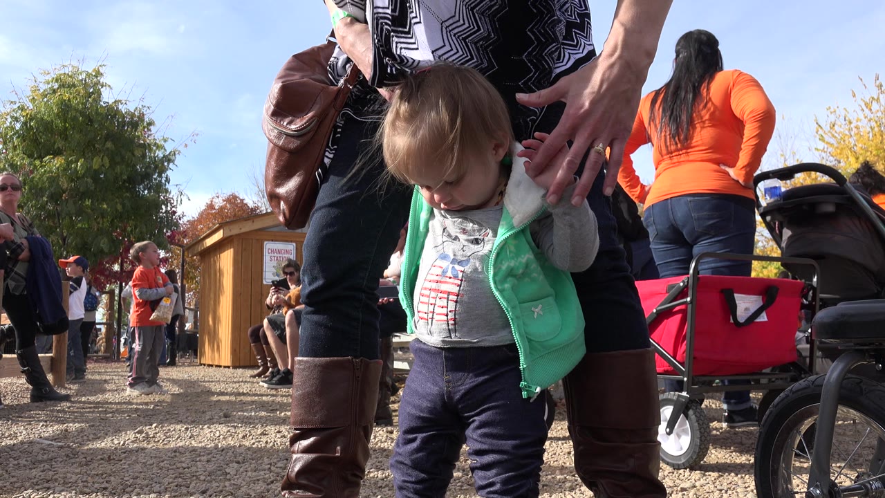 At 0:00 in Brighton, Colorado, a toddler in a mint vest looks down while an adult holds both hands, with a red wagon and park visitors behind them.