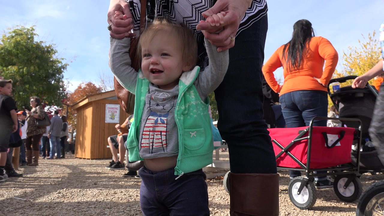 A toddler in a mint vest smiles while an adult holds her hands at a Brighton, Colorado outdoor fair, with fairgoers and a red wagon behind her.