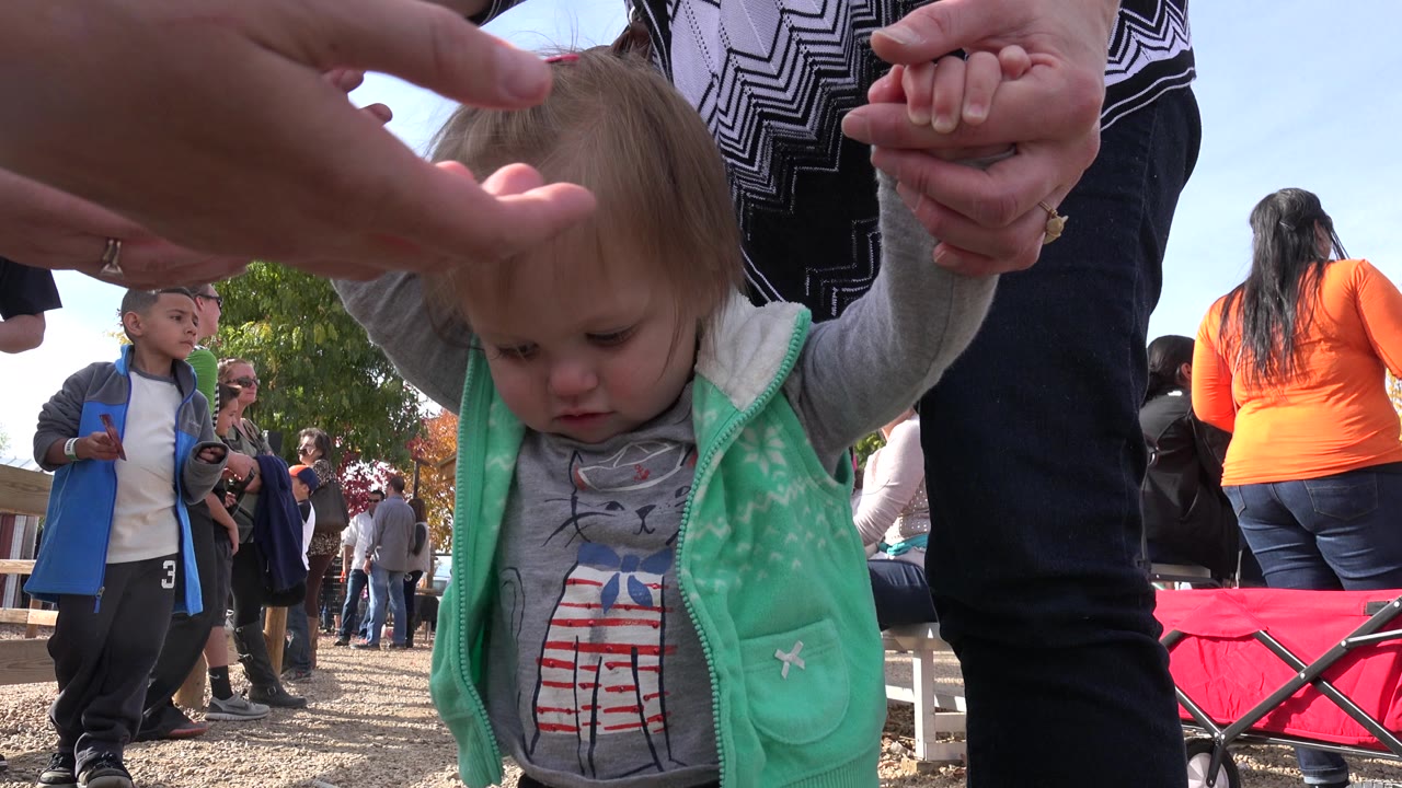 A blonde toddler in a mint green vest looks down while adult hands hold both arms at an outdoor gathering in Brighton, Colorado.