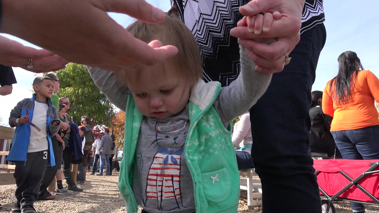 The toddler remains centered and close to the lens, with adult hands above her and a child in a blue vest visible behind her in Brighton, Colorado.