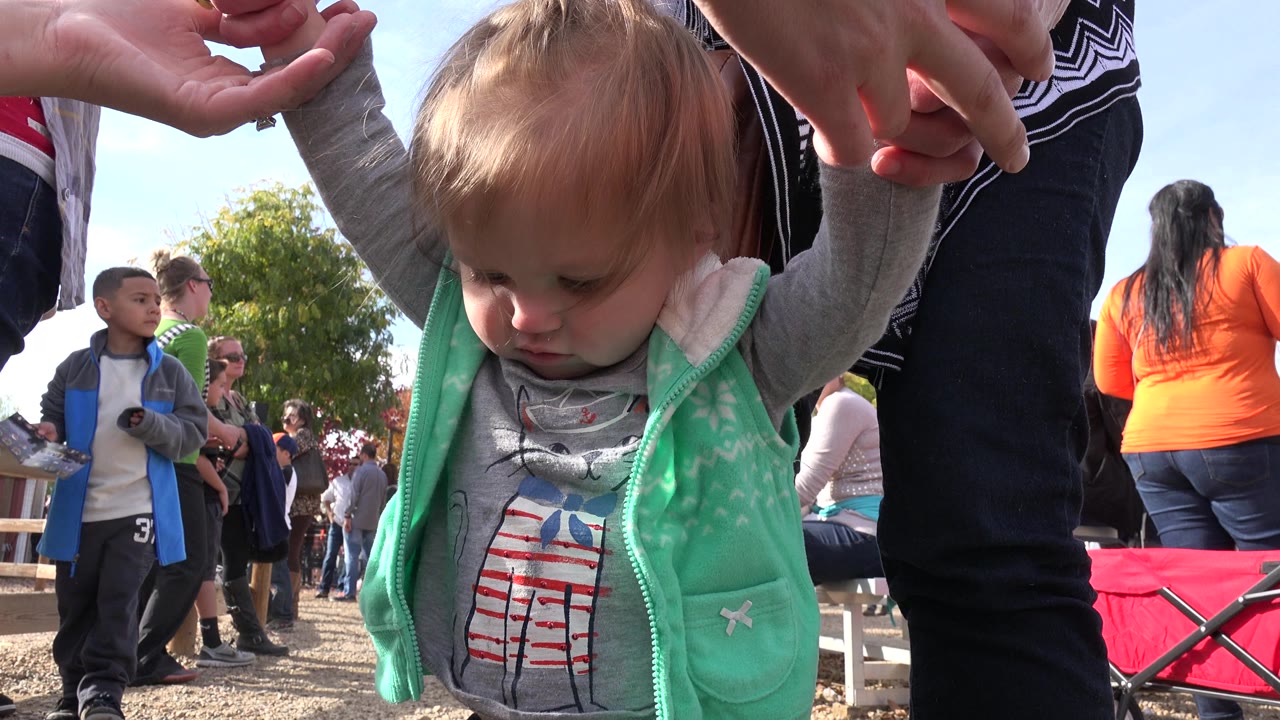 Toddler held upright with people and a red wagon behind her.
