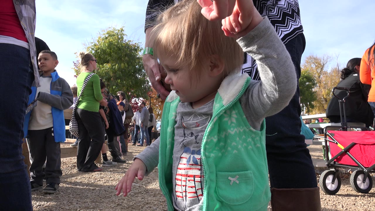 Toddler girl in a mint vest standing among adults at an outdoor gathering.