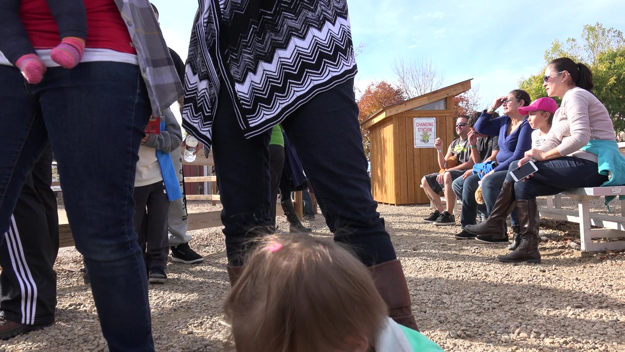 Adult legs fill the foreground in Brighton, Colorado, while several people sit on a bench near a wooden building under clear daylight.