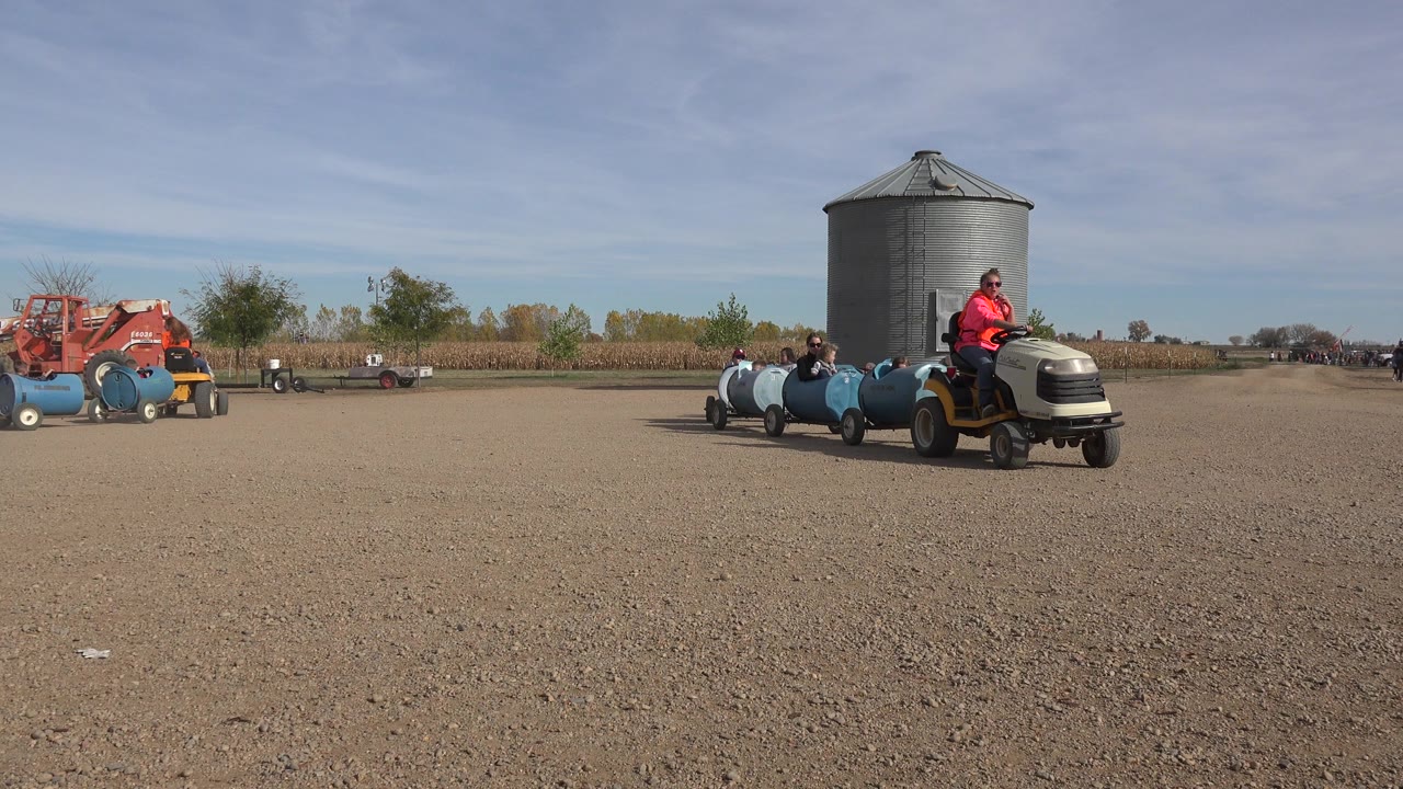 The tractor train moves nearer in Brighton, Colorado, showing the orange-shirted driver and several passengers riding in blue barrel cars beside the silo.