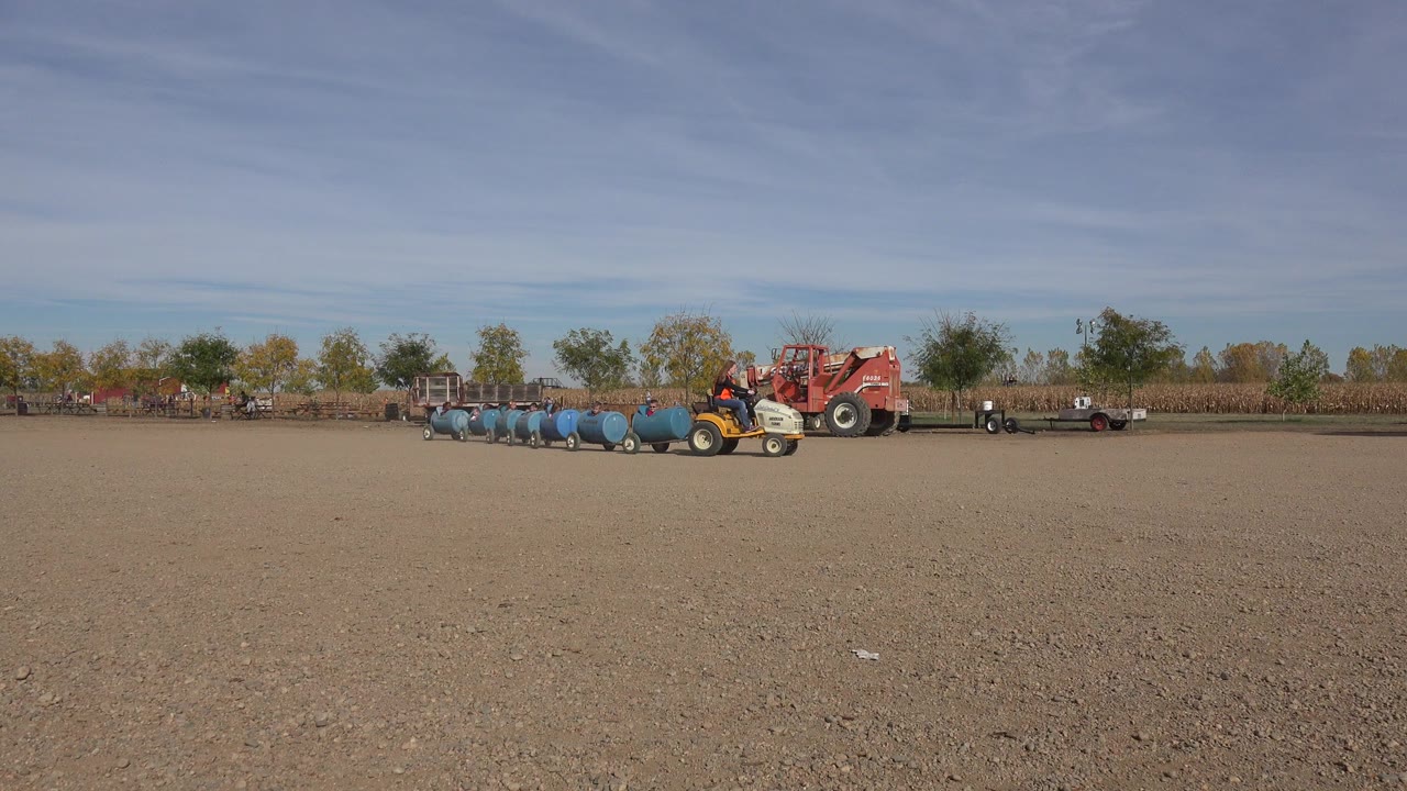 In Brighton, Colorado, a small yellow tractor pulls blue barrel cars across a broad dirt farm lot, with a red tractor, trees, and crop rows in the background.