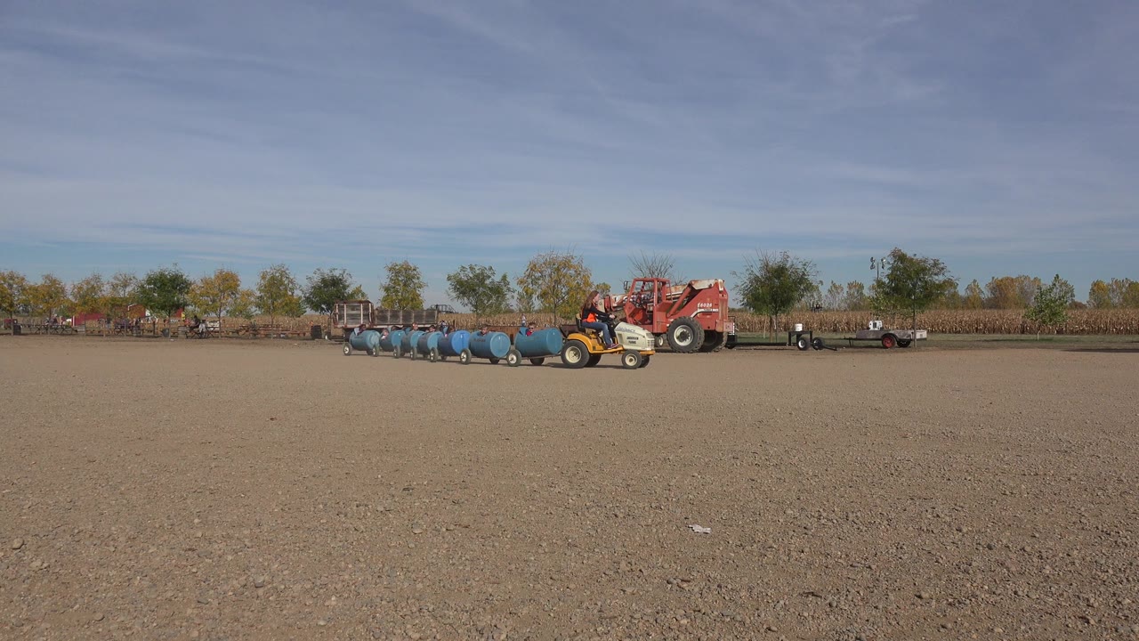The Brighton farm scene shows the barrel cars lined behind the small tractor near a red agricultural vehicle, with open dirt foreground and a low tree line beyond.