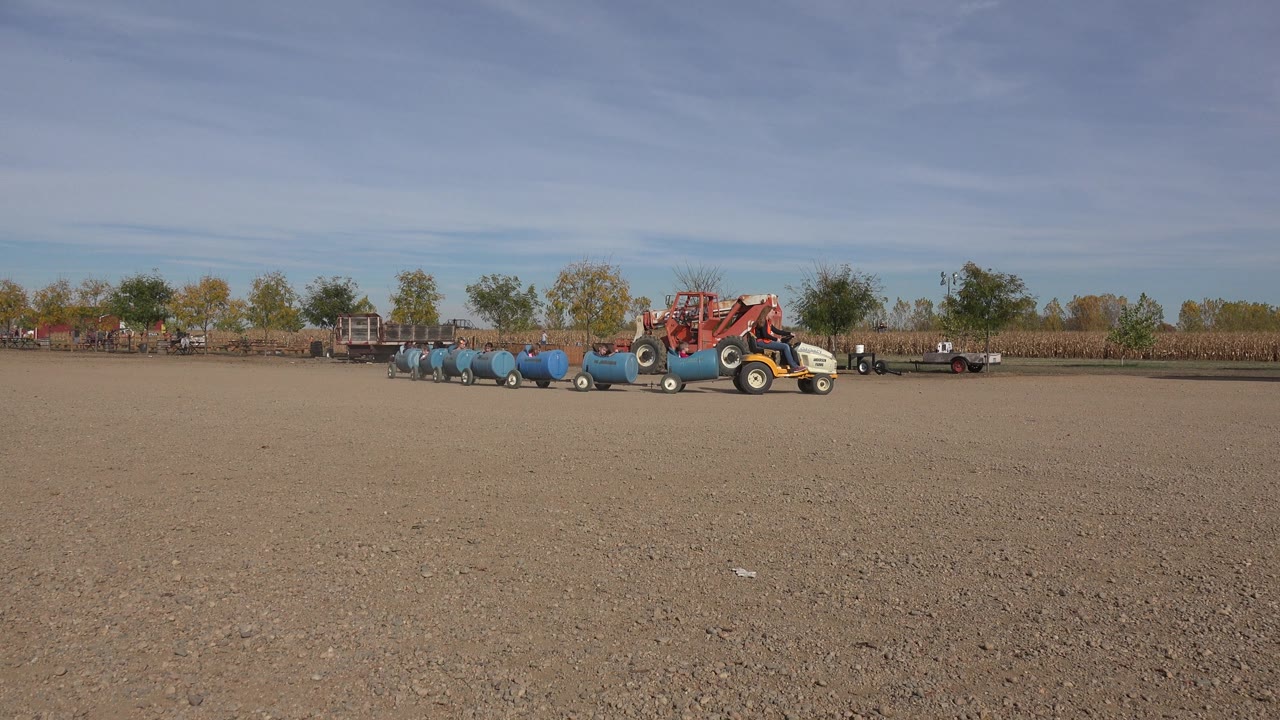 Barrel train and tractors moving across a farm lot under a bright sky.