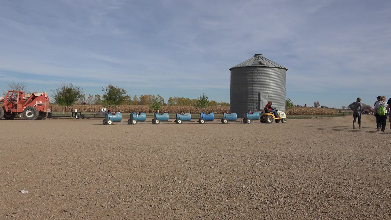 Wide field with grain bin, red tractor, blue train cars, and people.