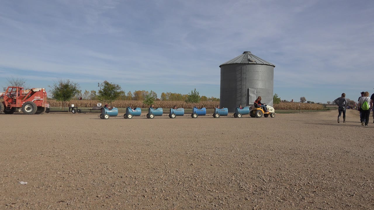 In Brighton, Colorado, two people walk through the open dirt foreground while the red tractor, gray grain bin, and blue train cars sit across the midground.