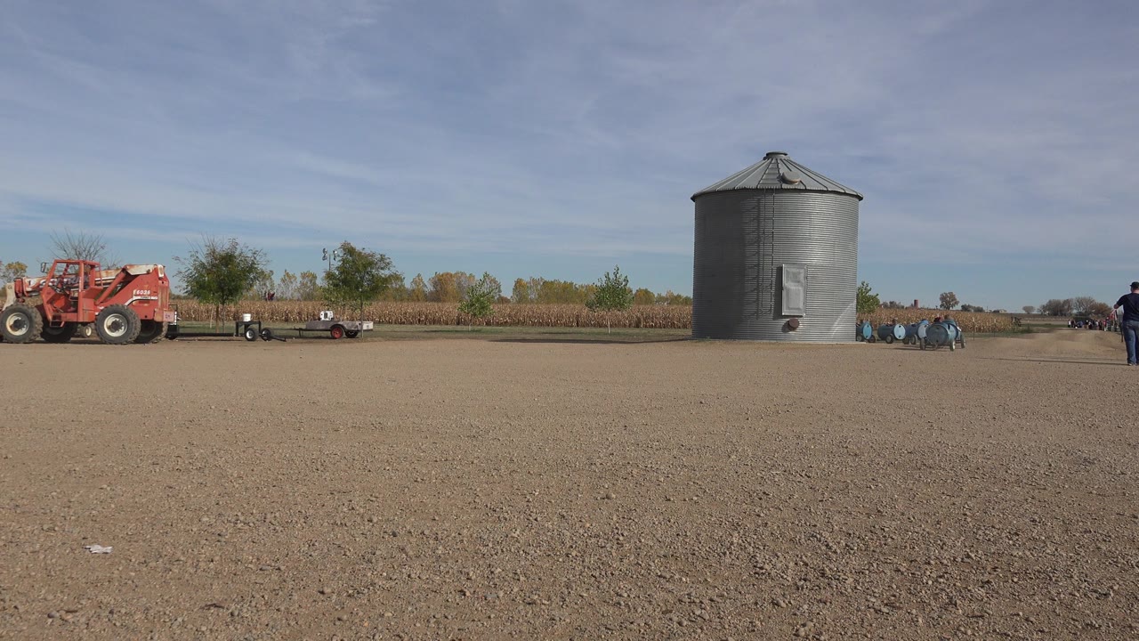Wide dirt farmyard with red tractor, grain bin, trailers, and a person standing nearby.
