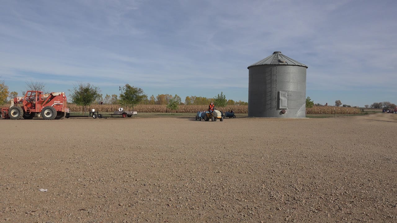 At the start of the Brighton, Colorado clip, a small tractor towing blue tanks moves across an open dirt field with a gray grain bin and red farm vehicle in the background.