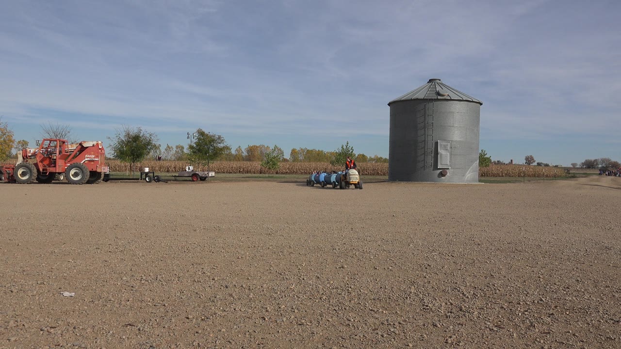 Small tractor and blue tanks pass to the right of a red farm vehicle.