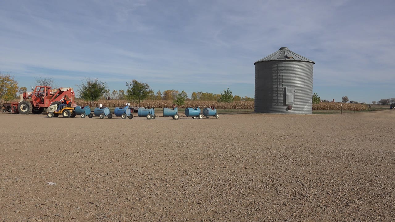 Wide view in Brighton, Colorado showing a dirt lot, red farm equipment, blue wheeled carts or barrels, a gray grain bin, distant trees, and no visible people.