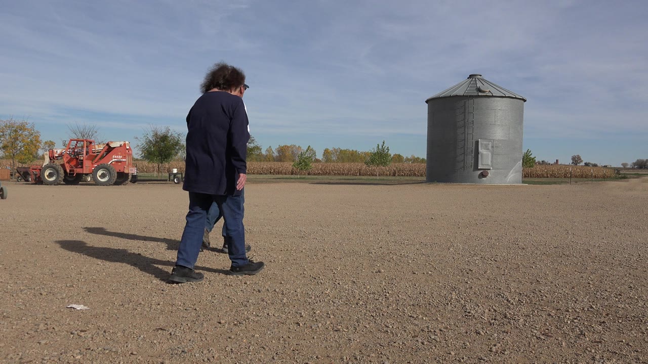 Man walking across a gravel lot near a grain bin and red equipment.