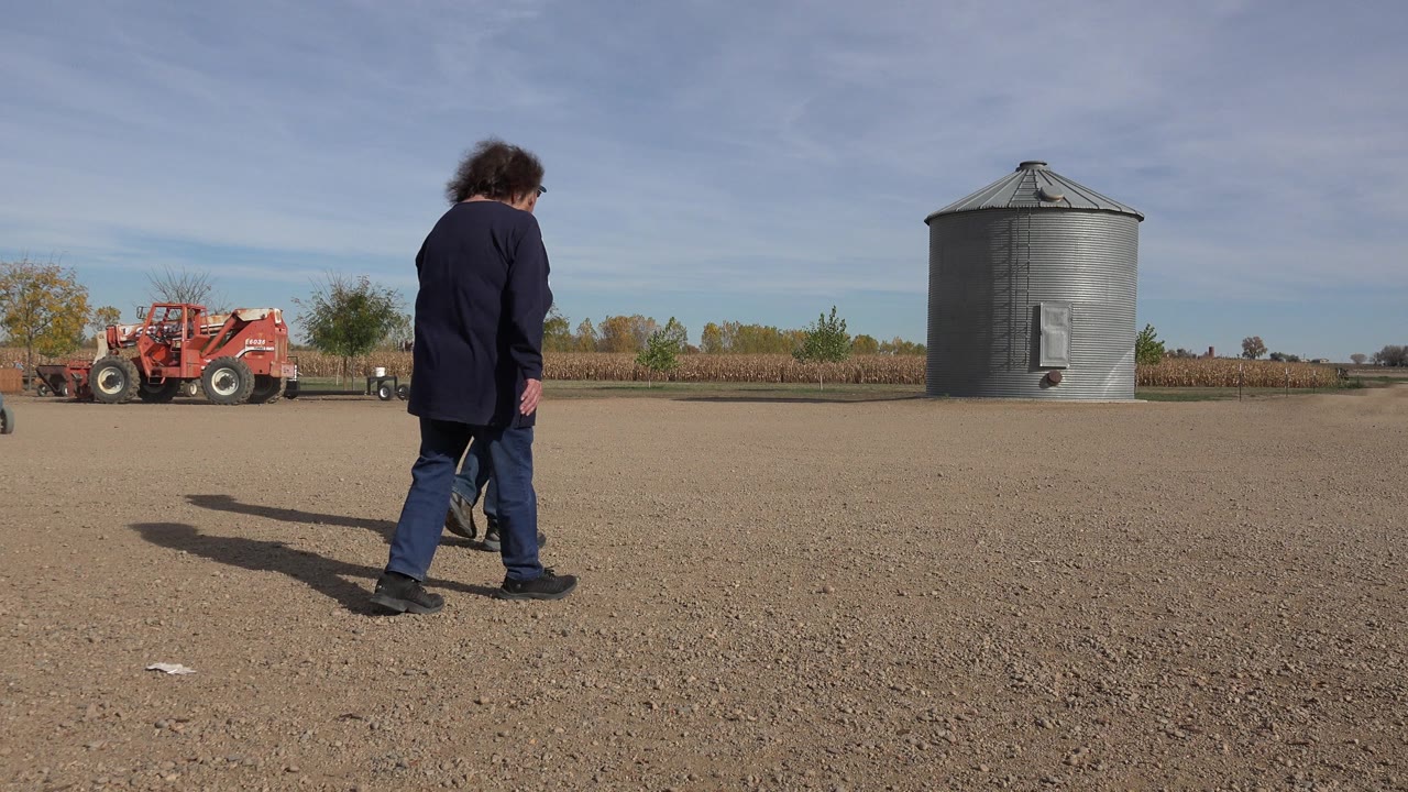 One man continues across the open gravel in Brighton, Colorado, with the grain bin to the right and red equipment near the left background.